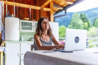 Happy freelancer woman working on laptop from the balcony of a mountain chalet, enjoying freedom