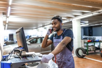 Young mechanic wearing gray uniform and protective glasses using computer and talking on phone