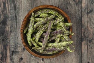 A wooden bowl filled to the brim with vibrant purple and green beans rests on a textured wooden