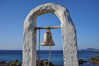 Close-up of a bell in a white frame in front of a rocky coastal landscape and blue sky, rock church