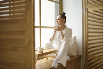 Dreamy woman in white clothes sitting on windowsill and holding cup of coffee or tea with both