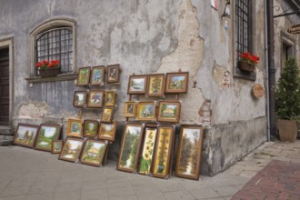 Framed paintings displayed against an old damaged grey cement wall in the Old Town Market Square,