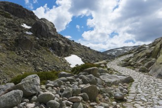 A stony path through a rocky mountain region with patches of snow under a cloudy sky, hiking area,