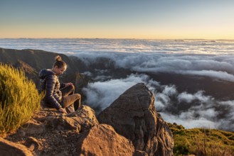 A woman sits on a rock and looks at the sea of clouds below her at dusk, Pico Ruivo, Madeira,