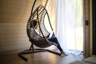 On a hot sunny day, a young woman sits in a hanging swing chair with a laptop in an air-conditioned