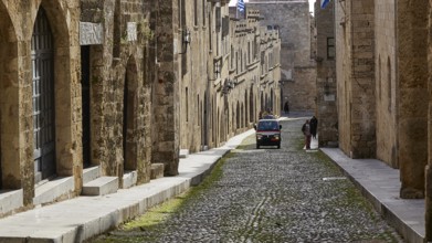 A historic cobblestone street with old yellow stone buildings, a moving car and a pedestrian,