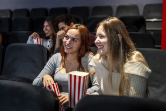 Two young women enjoying popcorn and smiling while watching a movie together in a cozy cinema,