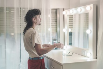 Young man with a mullet hairstyle standing in front of a lit mirror, holding items, dressed in a
