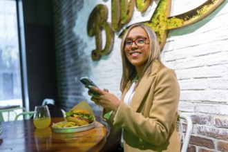 Latin woman smiling at camera sitting on a restaurant of fast food eating cheeseburger while using
