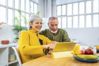 Happy senior couple using digital tablet at home pointing at the screen and smiling while eating