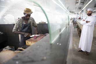 Omani man filleting a fish in Muttrah fish market, an Omani man in dishdasha watches, Muscat, Oman,