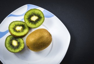 One whole kiwi and two cut kiwis on a white plate with a black background