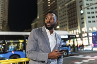 Young businessman holding a smartphone, looking around in a bustling city at night, surrounded by