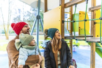 Cheerful parents and their baby are enjoying a playful day at the playground during wintertime
