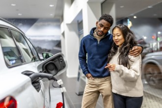Multiethnic couple examining charging port of electric vehicle at car dealership, embracing