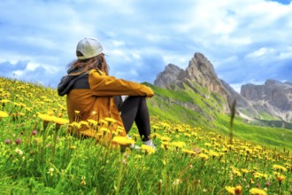 Young woman sitting in a field of yellow flowers admiring the odle mountain group in the italian