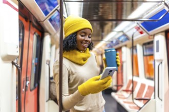 Smiling woman wearing yellow winter clothes using phone and holding reusable coffee cup while