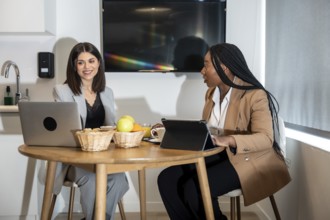 Two businesswomen are enjoying breakfast and working with laptop and tablet in their hotel room