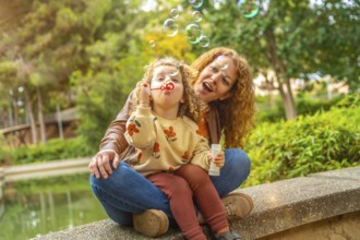 Caucasian happy mother and girl blowing soap bubbles sitting in the park