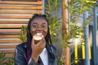 African cute woman eating a colorful doughnut sitting outdoors next to a shop