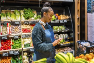 Smiling saleswoman wearing apron using digital tablet and managing inventory of fresh organic