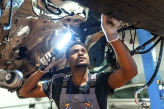 Young mechanic using flashlight, inspecting underside of vehicle raised on lift in professional