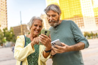 Senior smiling woman showing mobile screen to senior partner outdoors in the city