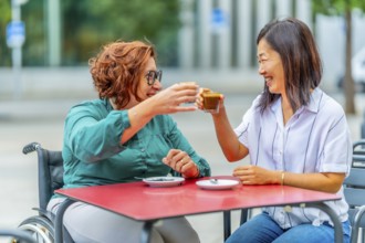 Woman with disability and asian friend toasting with coffee sitting on an outdoor cafeteria in the