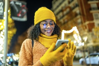 Smiling young woman wearing yellow winter clothes and using smartphone in a city street at night