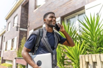 African man carrying laptop and using mobile in the street walking along a residential area