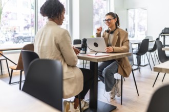 Two women engaging in a podcast recording session at a cafe, utilizing a laptop and microphone for