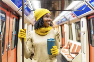 Smiling young woman wearing yellow winter clothes and holding reusable coffee cup standing inside