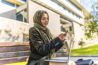 Muslim businesswoman using mobile working on remote with a laptop sitting on a bench in a park