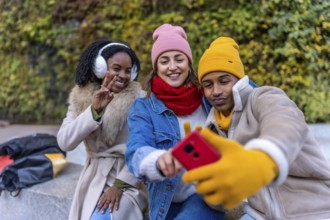 Three happy young students are taking a selfie in a city park during winter, wearing warm colorful