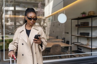 Stylish businesswoman using smartphone while holding luggage outside hotel during business trip,