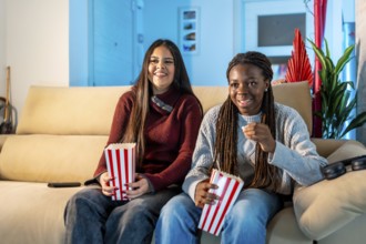 Two happy teenage girls eating popcorn while watching a movie at home, sharing a moment of leisure