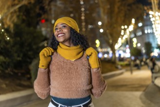 Smiling tourist wearing winter clothes looking up at christmas lights in a city center at night