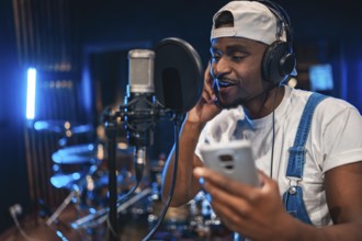 Young man recording vocals in professional music studio using smartphone, microphone and headphones