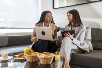 Two businesswomen discussing work on a business trip, using laptop and tablet in hotel room