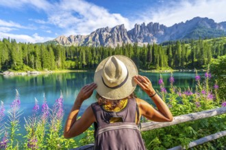 Woman with backpack and straw hat admiring the turquoise waters of lake carezza and the latemar