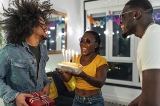Young multiracial people surprising a friend with birthday cake and gift during night party at home