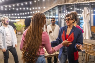 Two multi-ethnic friends women dancing together next to other friends at night party outdoors