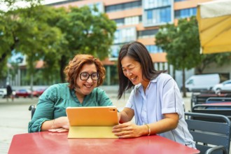 Disabled woman in electric wheelchair and chinese female friend using tablet sitting on sidewalk