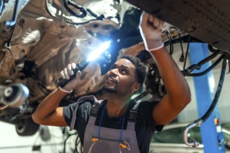 Young mechanic using flashlight, inspecting underside of vehicle raised on lift in car workshop