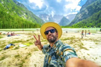 Tourist wearing sunglasses and straw hat gesturing peace sign while taking a selfie at the beach of