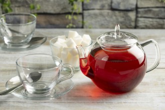 A teapot with red tea next to empty glass cups and sugar cubes on a table against a stone