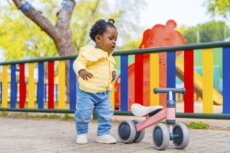 Full length photo of an african baby girl in a playground with a toy bike alone