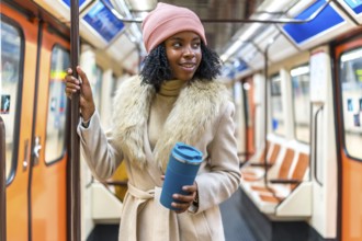 Stylish young woman holding a reusable coffee cup while standing on a subway train, enjoying her