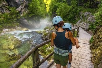 Mother carrying her child while walking on a wooden pathway, exploring the scenic beauty of vintgar