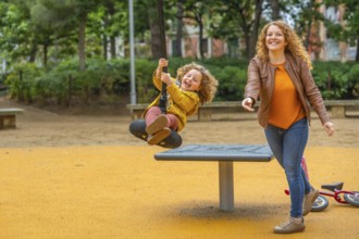 Caucasian adult mother and girl having fun swinging in a playground in an urban park